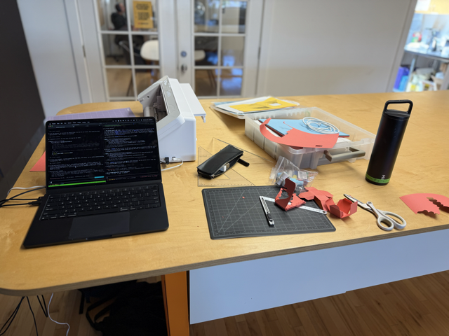 Photo of a large desk, with a laptop, a Silhouette Cameo cutter. The laptop has a split screenw with two terminals